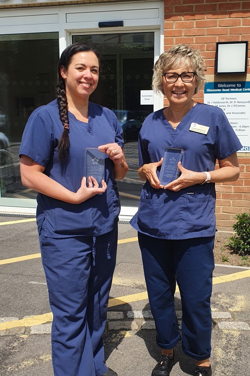 Image shows two female nurses standing in front of the practice building holding this awards